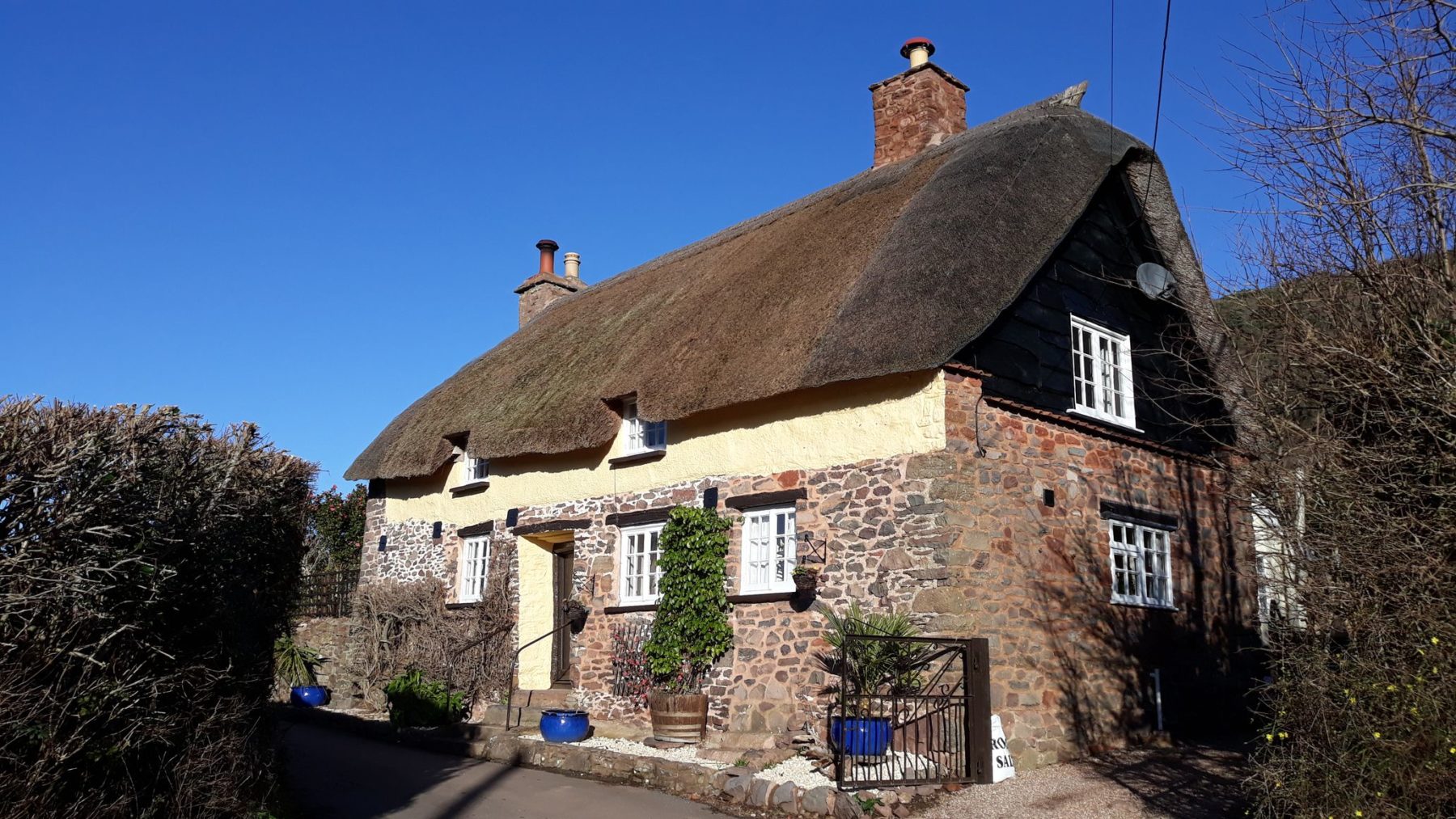 A photo of Jasmine Cottage, Allerford - a picturesque two-storey cottage with rubble stone walls, limewash, and a thatched roof.