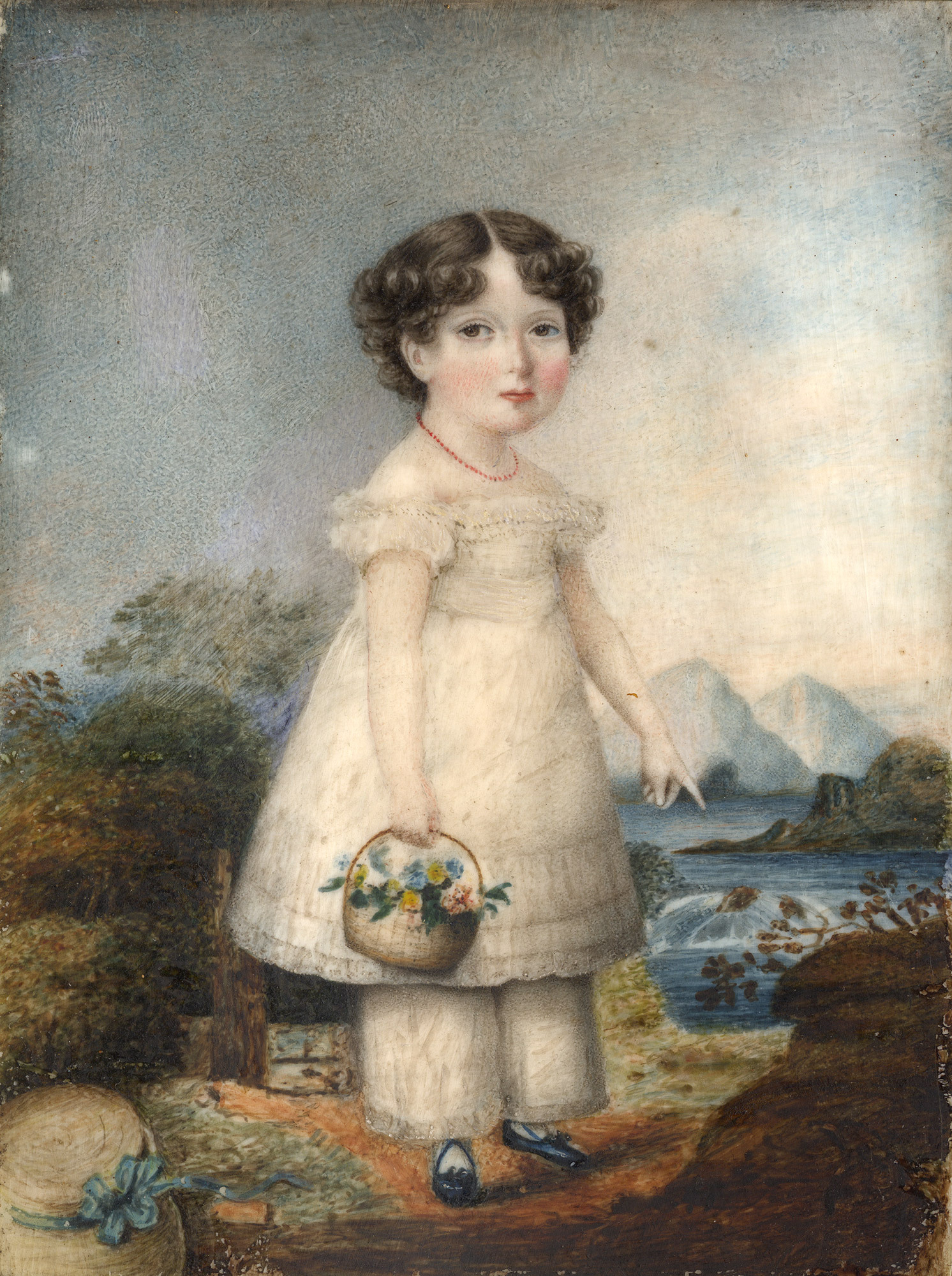 Portrait of a young girl, standing, wearing a white dress, pantaloons, black shoes and a coral necklace. She holds a basket of flowers in her near hand, and is against a backdrop of water and trees.