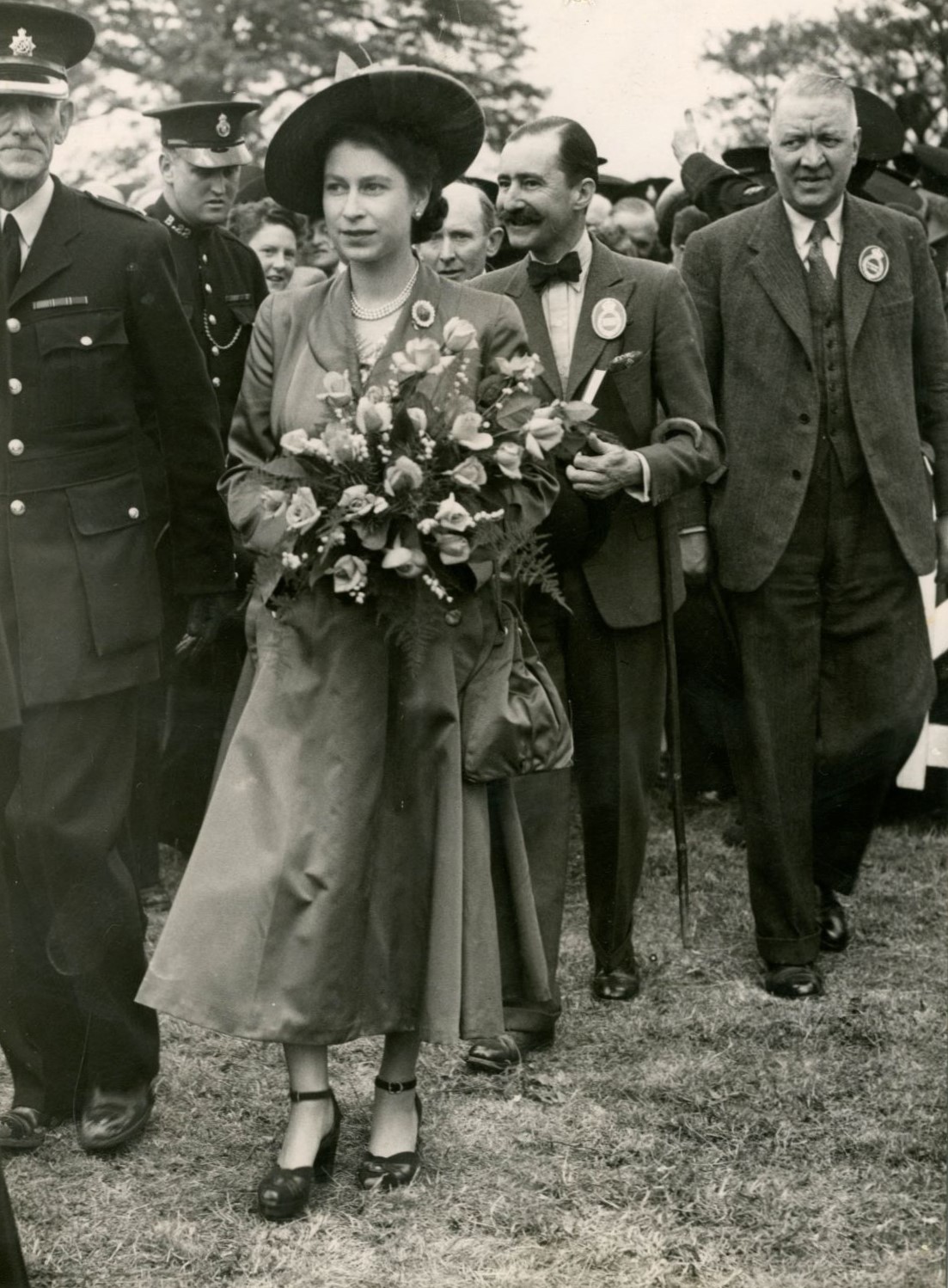 Black and white photograph of young Queen Elizabeth II walking, holding a bouquet of flowers, with people walking beside and behind her.