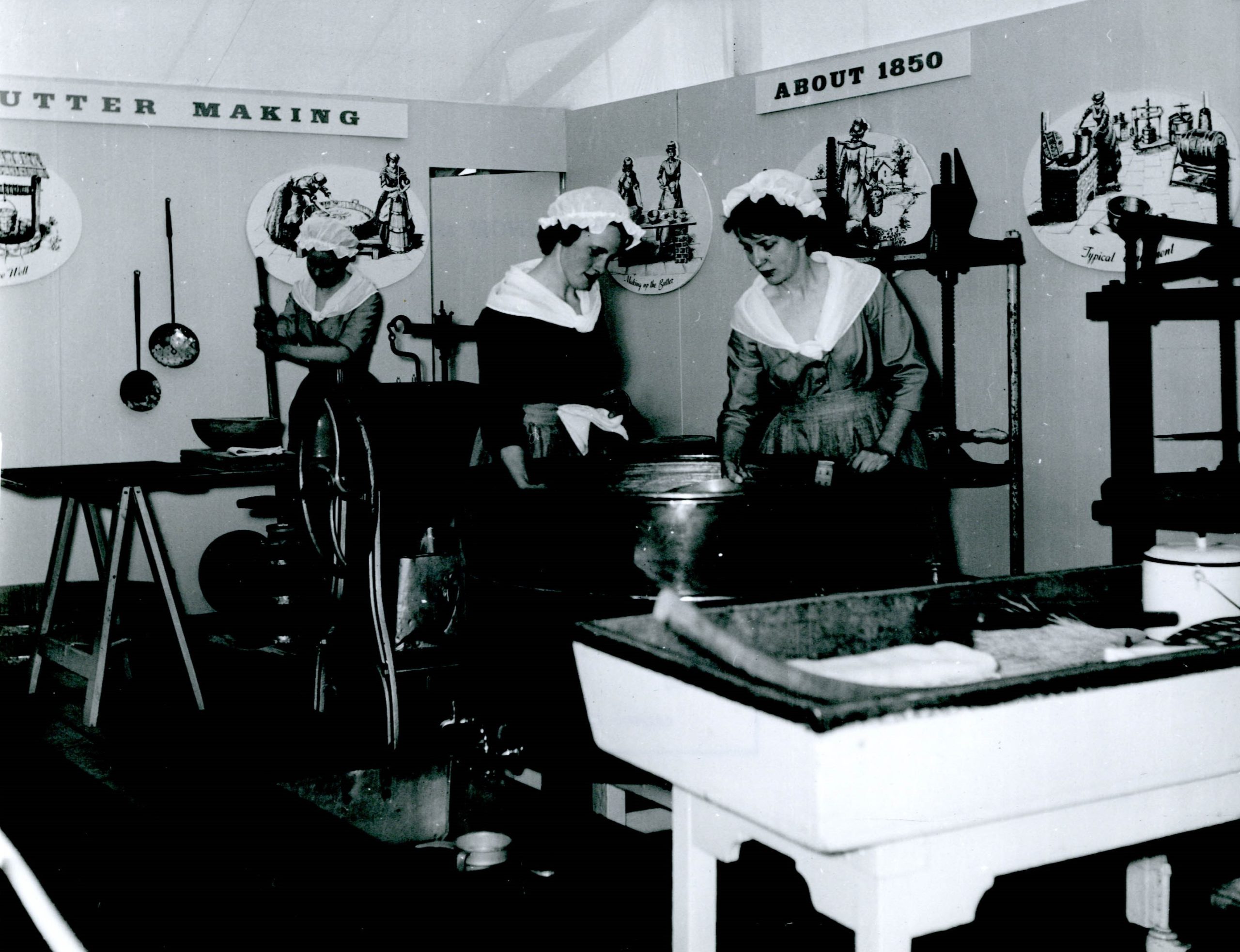 Black and white photograph showing three women dressed in aprons with butter churns and other equipment around them. They are in a Show tent, with a display sign reading 'Buttermaking, About 1850s'.