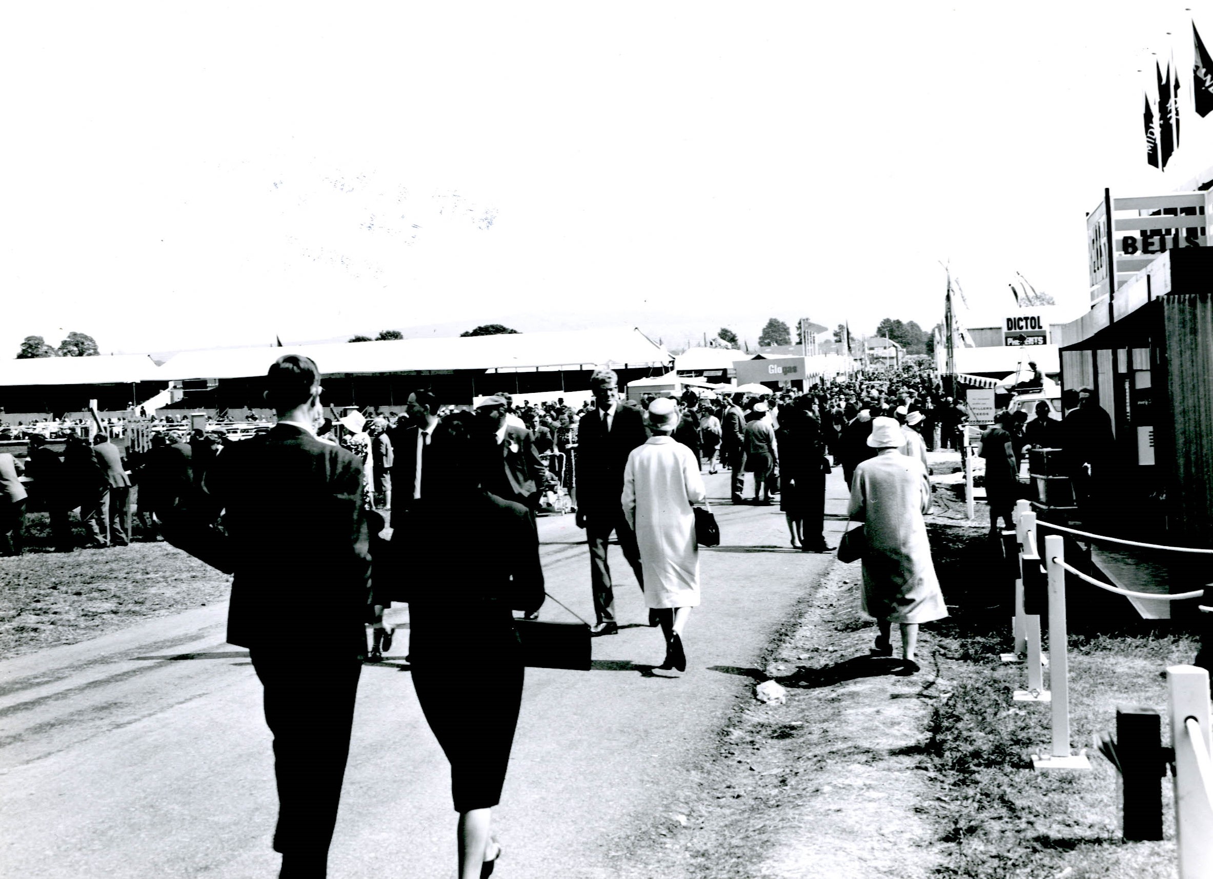 Black and white photograph of a busy crowd of people with the main ring grandstand partially visible above and to the left. Trade stands line the right hand side.