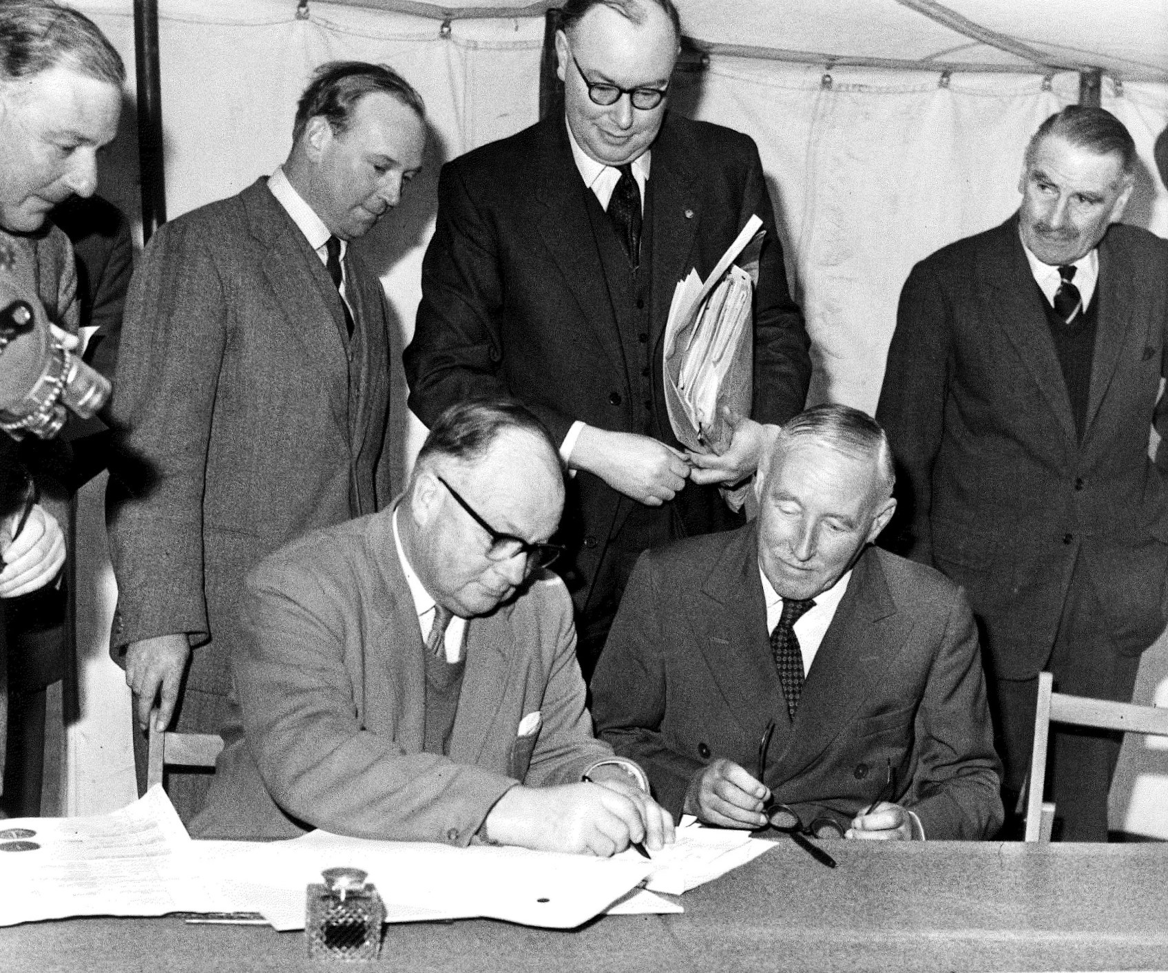 Black and white photograph of 2 men sat at a table facing the camera. They are signing papers as 4 other men look on from behind.