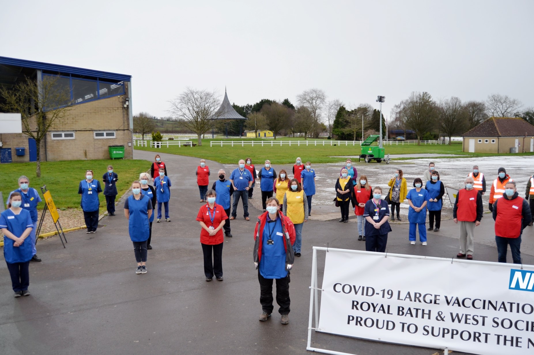 Colour photograph of vaccination centre staff fanned out with face masks on. They stand facing the camera, with Shepton Mallet Showground as the backdrop. A sign is partially obscured on the right, it reads 'Covid-19 large vaccination centre, Royal Bath & West Society, proud to support the NHS.'