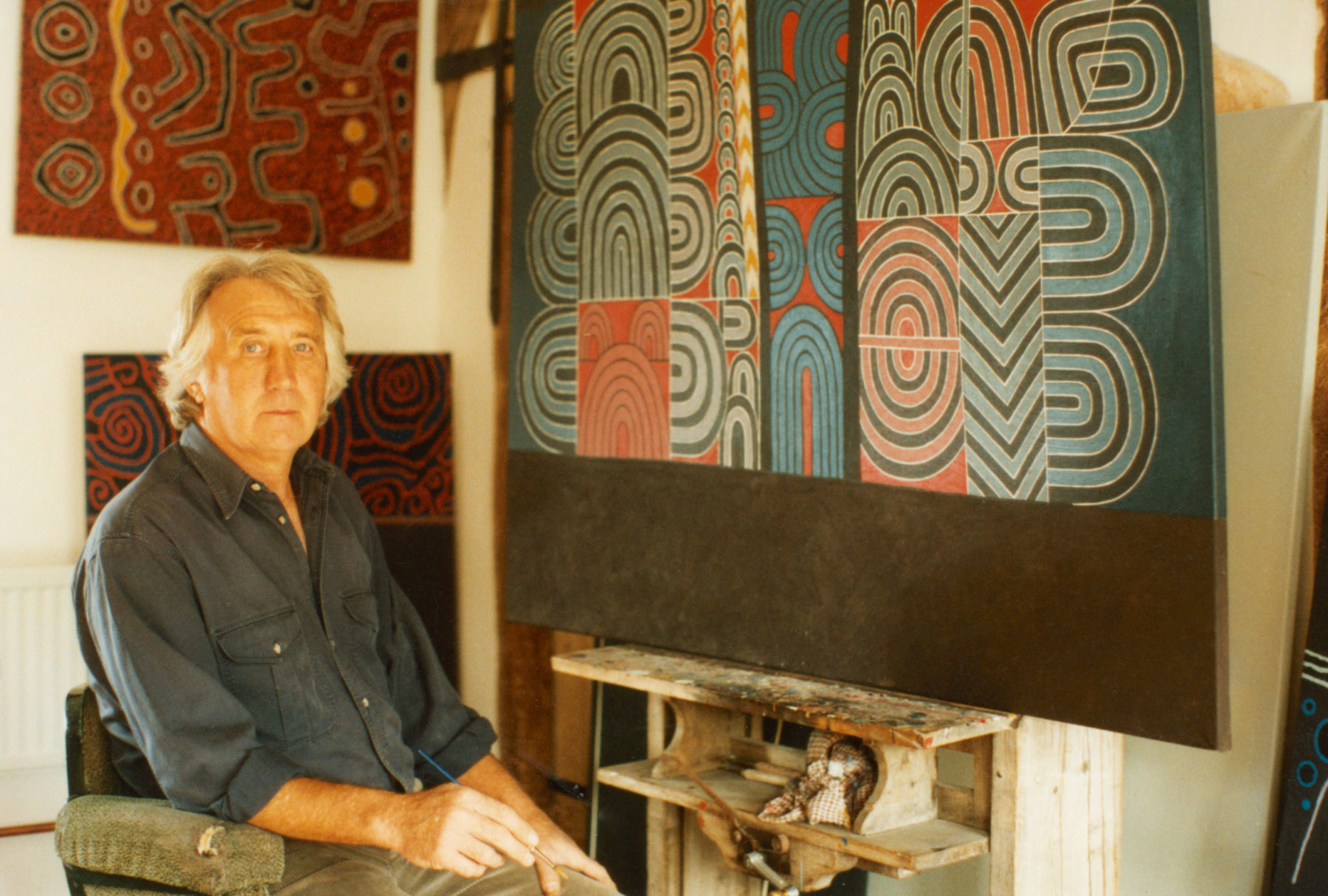 Colour photograph of Brian Rice sat in a chair alongside several of his works, which are hanging on the wall. He is holding a paintbrush and appears to be in a studio.
