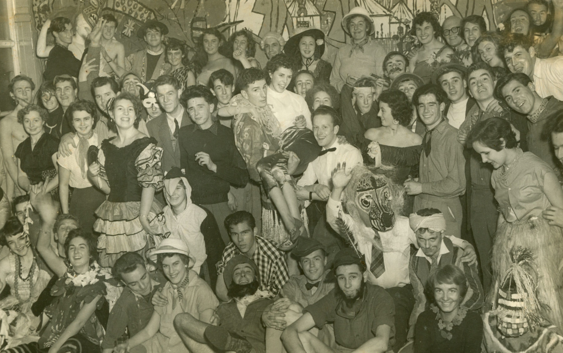 Black and white photograph of a large group of students, smiling and posing for the camera. The crowd wear various formal wear and novelty costume.