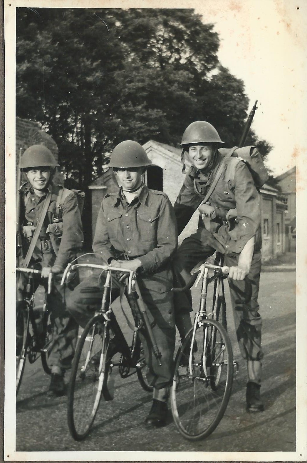 Black and white photograph depicting young Brian Rice, centre, cycling towards the shot with two friends cycling either side. All three are wearing army uniforms, helmets, and have backpacks and firearms strapped to their backs. They are smiling at the camera.