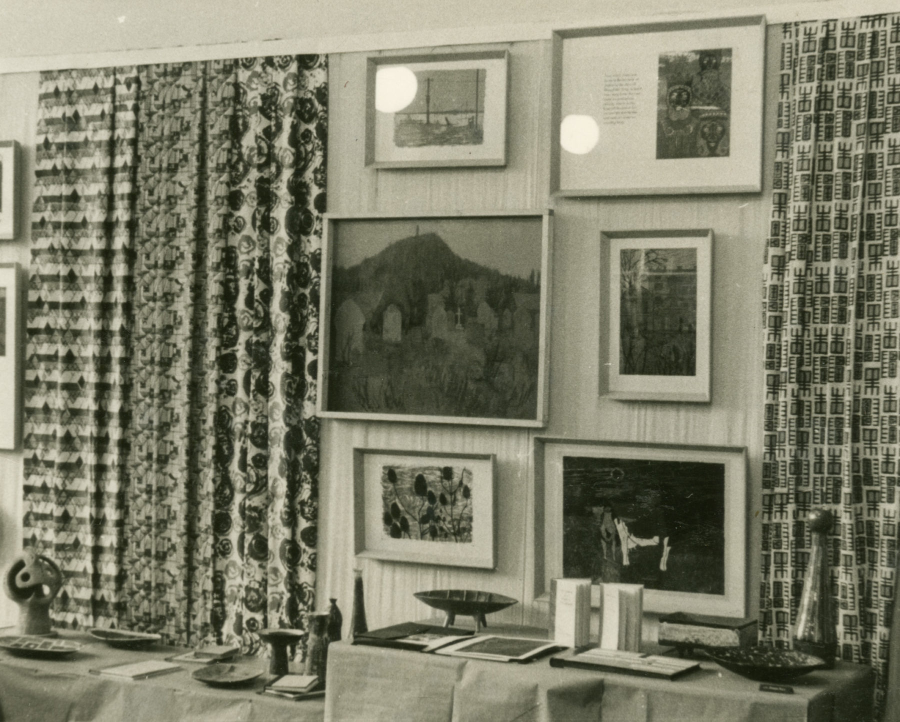 Black and white photograph of artworks mounted on a wall in front of a dressed table. Six works are hanging in two columns on the wall, the middle of which is pictured the framed work, St Michael's Mount from Churchyard.