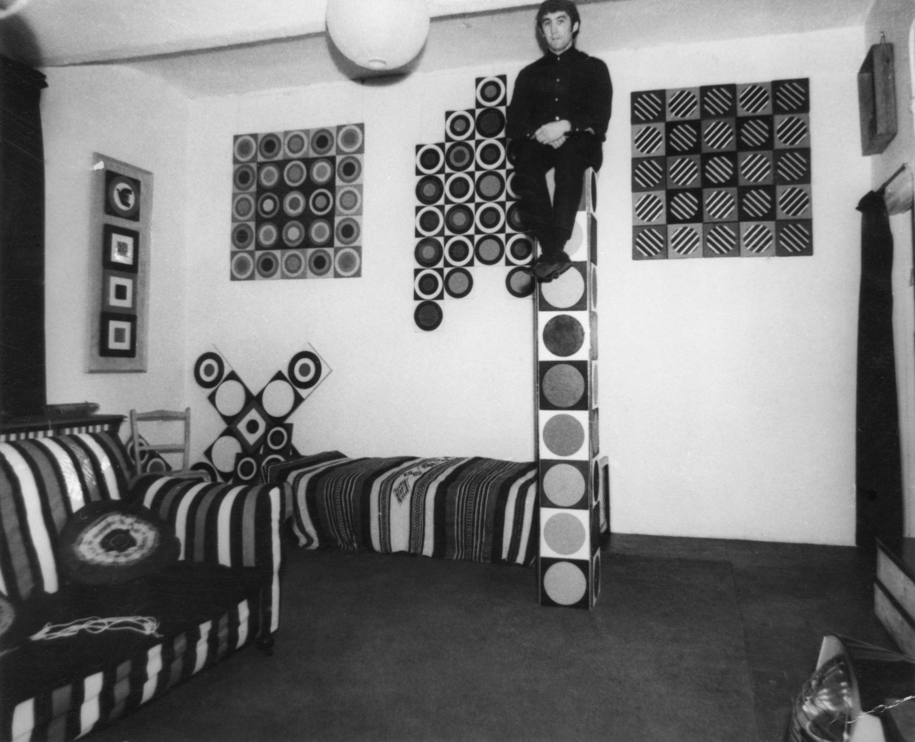 Black and white photograph of a small studio flat, with sofa and bed shown. Several of Brian Rice's paintings are displayed on the walls. Brian Rice himself can be seen sat atop a tall vertical pillar of his own making.