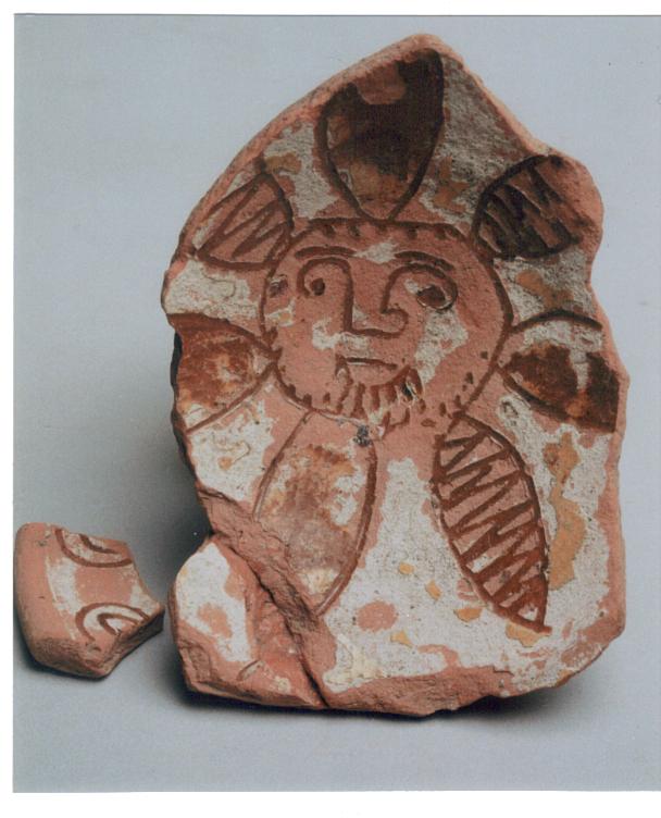 Photograph of a worn and cracked terracotta dish bearing a face framed by petal shapes. A broken fragment is displayed next to the dish.
