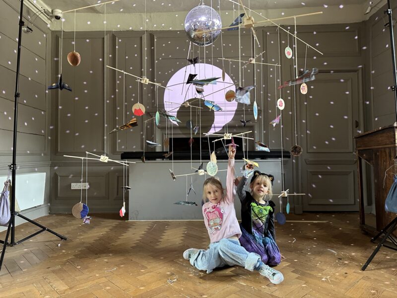 Two children sat underneath a large, outer-space themed mobile.