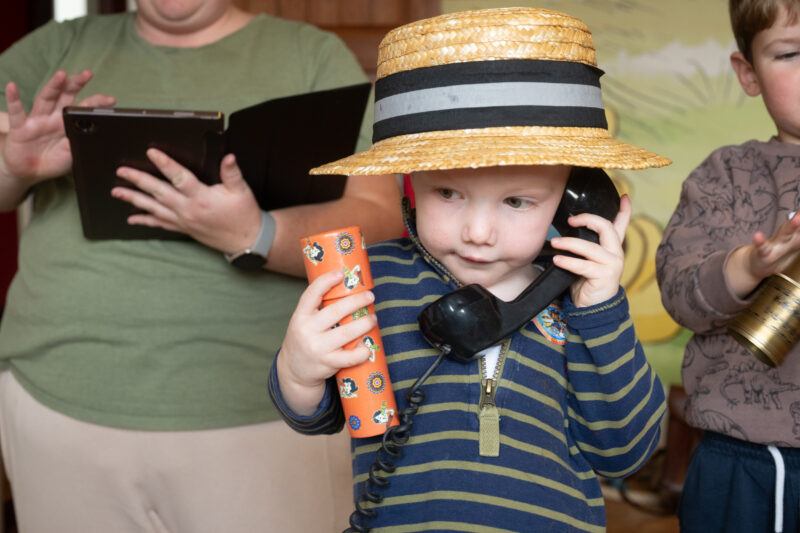Child wearing a hat and holding a telephone and a vintage toy, facing the camera.
