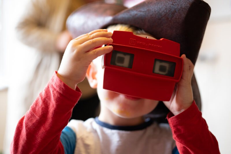 Child wearing a pirate hat looking towards camera through a viewmaster toy.