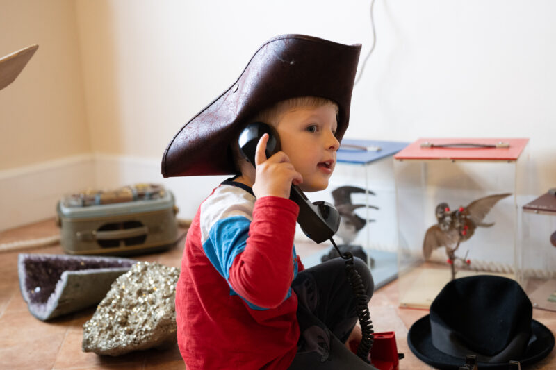Child wearing a pirate hat playing with a telephone, in front of boxes of taxidermy birds.