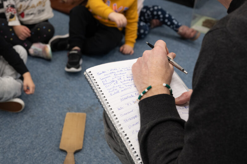 Man sat across from children, writing in notebook. Photo taken from over the man's shoulder.