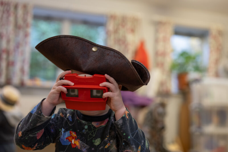 Child wearing a pirate hat, looking through a viewmaster toy at the camera.
