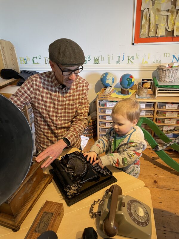 An adult sat next to a child playing with a vintage typewriter.