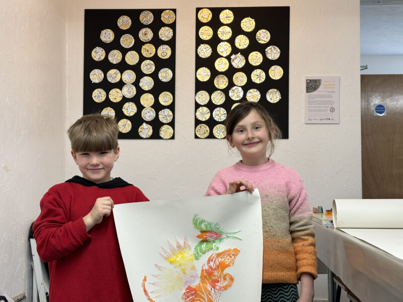 Two children holding artwork posing in front of display.