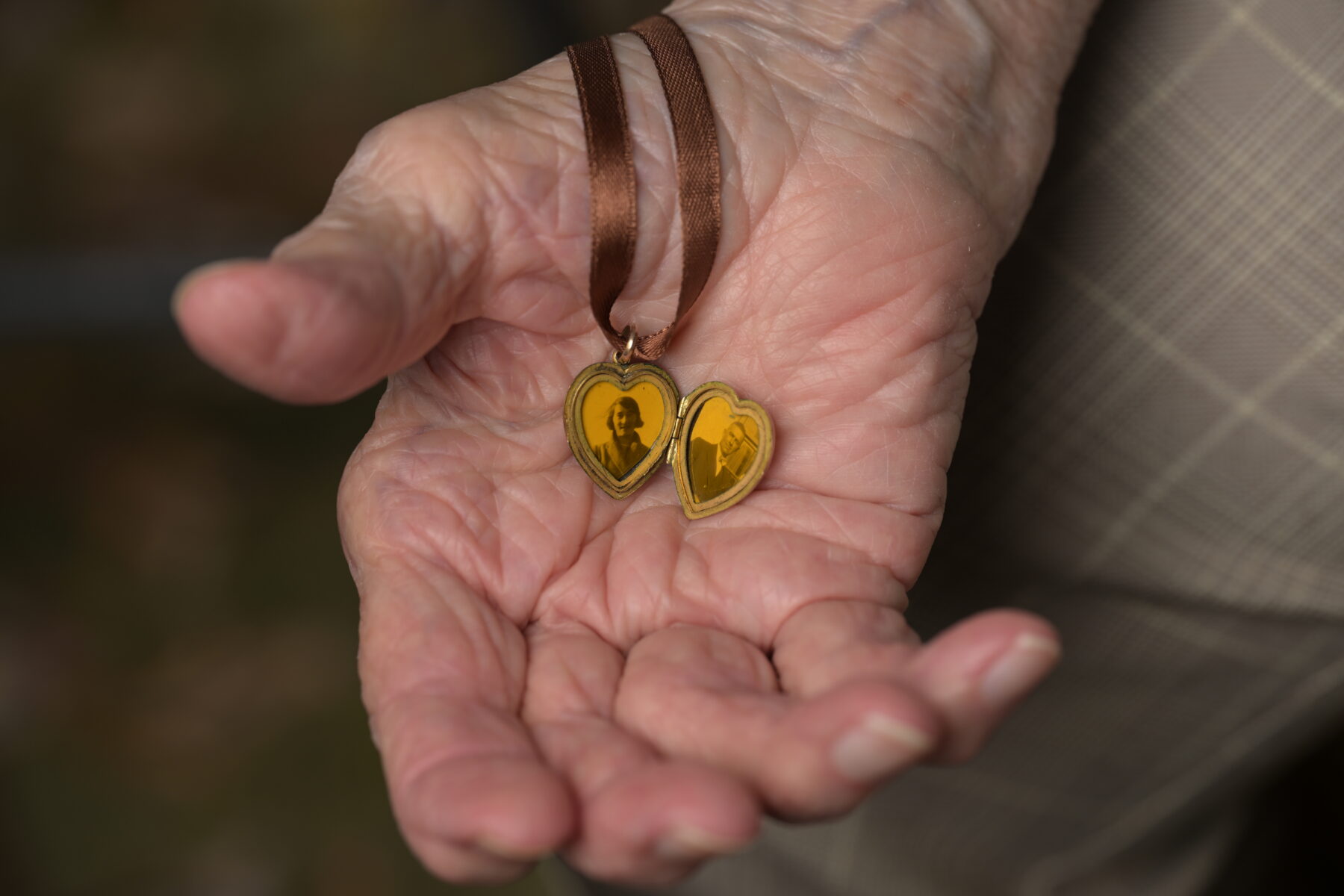 An elderly woman's hand holding a tiny heart shaped locket, showing a photograph of each of her parents.