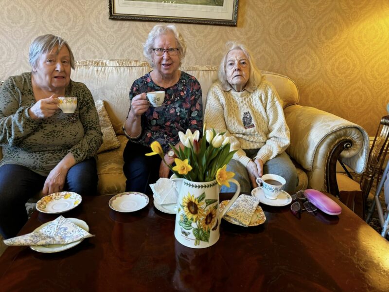 Three elderly women sat on a sofa holding cups of tea. Daffodils in the foreground.