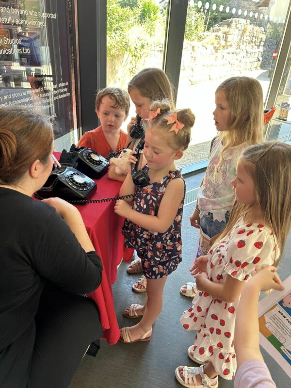 A crowd of children stood around a woman who is crouched down near some rotary telephones.