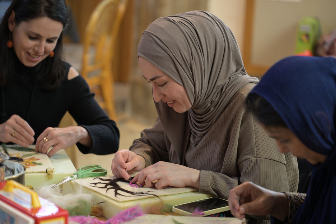 A woman wearing a grey hijab, smiling down at her artwork on the table. She has two women either side of her.