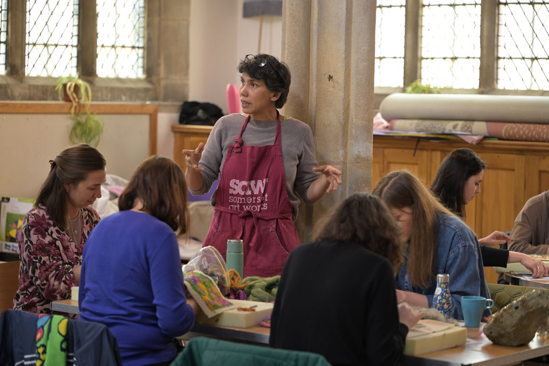 A woman with short hair and a red apron standing and talking to a group of women around a table.
