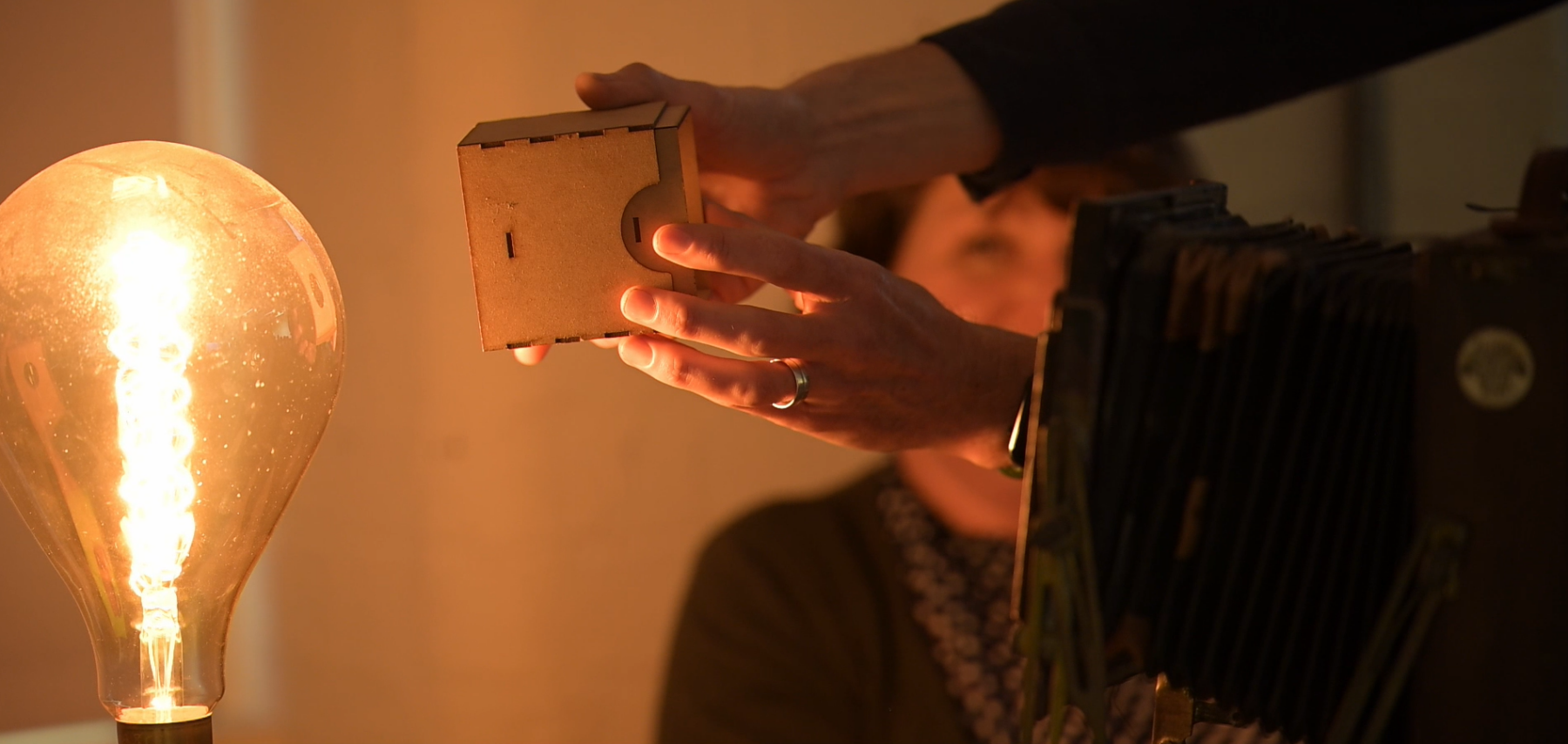 Hands holding a small homemade camera obscura between a vintage camera and a large lightbulb.