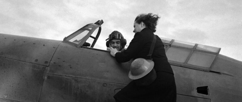 A young woman balances on the wing of a grounded aeroplane giving a message to the male pilot.