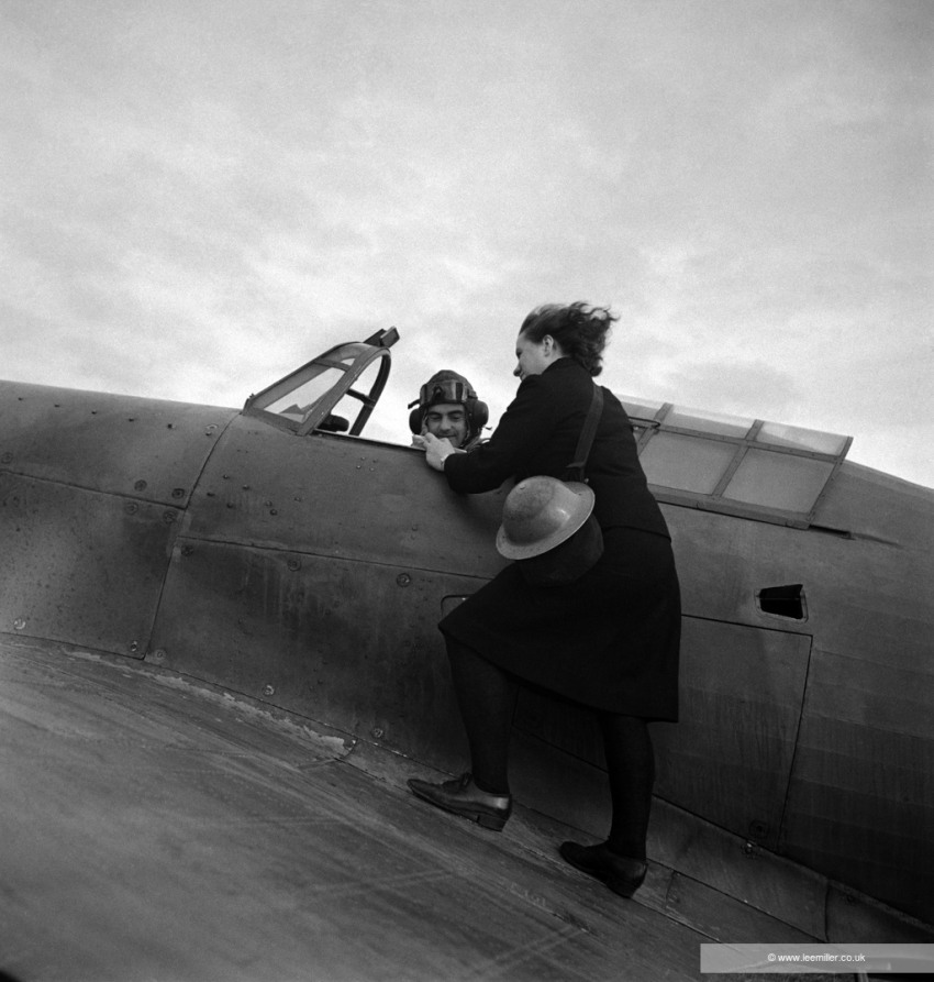 A young woman balances on the wing of a grounded aeroplane giving a message to the male pilot.