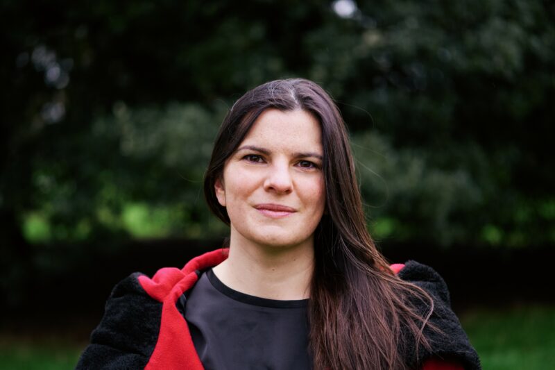 A headshot of the author of 'Cunning Folk: Life in the Era of Practical Magic'. She is stood in front of some trees and has long brown hair and brown eyes.
