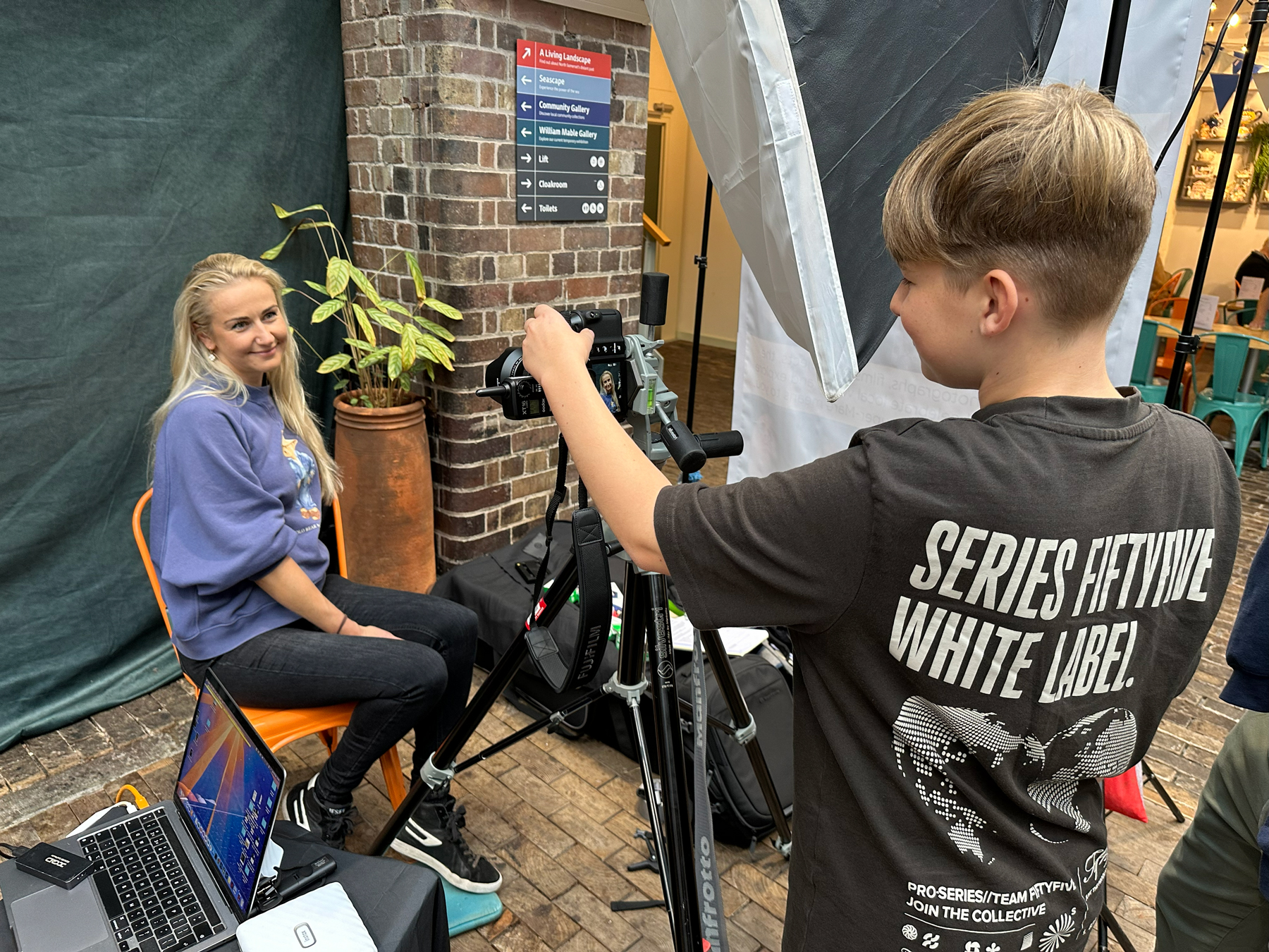 A young boy taking a photographic portrait of his mother, who is sat in front of a green background.