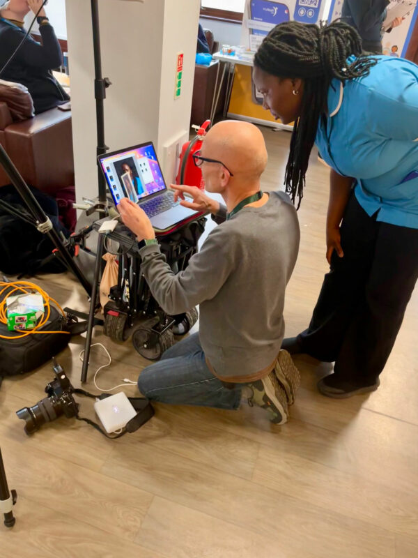 Man knelt down in front of a laptop, showing photographs with a woman in a blue NHS uniform looking over his shoulder.