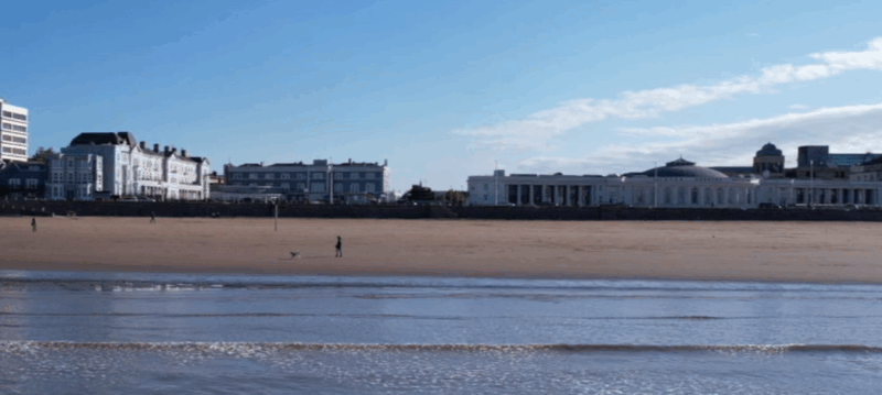 A photograph of small waves hitting a beach, with a dog walker and sea front buildings in the background.