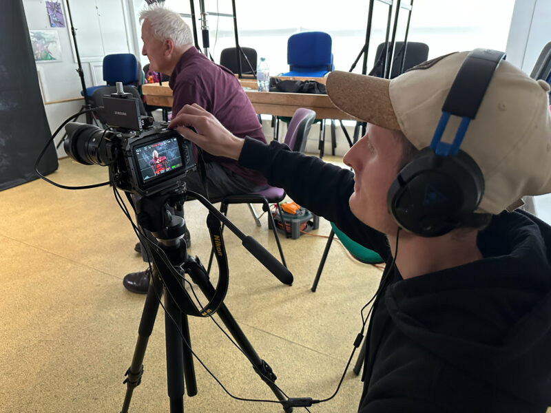 Man operating a camera in foreground, wearing a cap and headphones. Man in background interviewing a participant.
