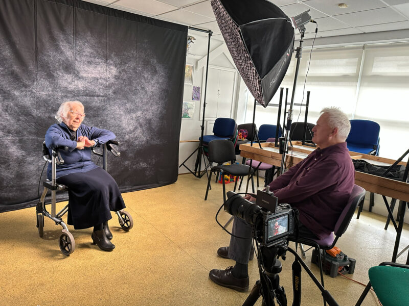 A pop-up filming studio, with a background and lighting set up. A man interviewing an elderly woman.