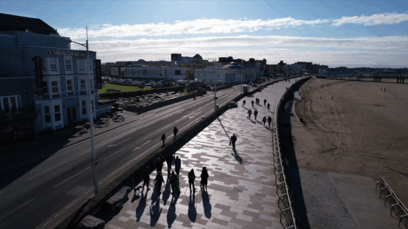 Groups of people walking along a sea front promenade, with a road and buildings on one side, and a beach on the other.