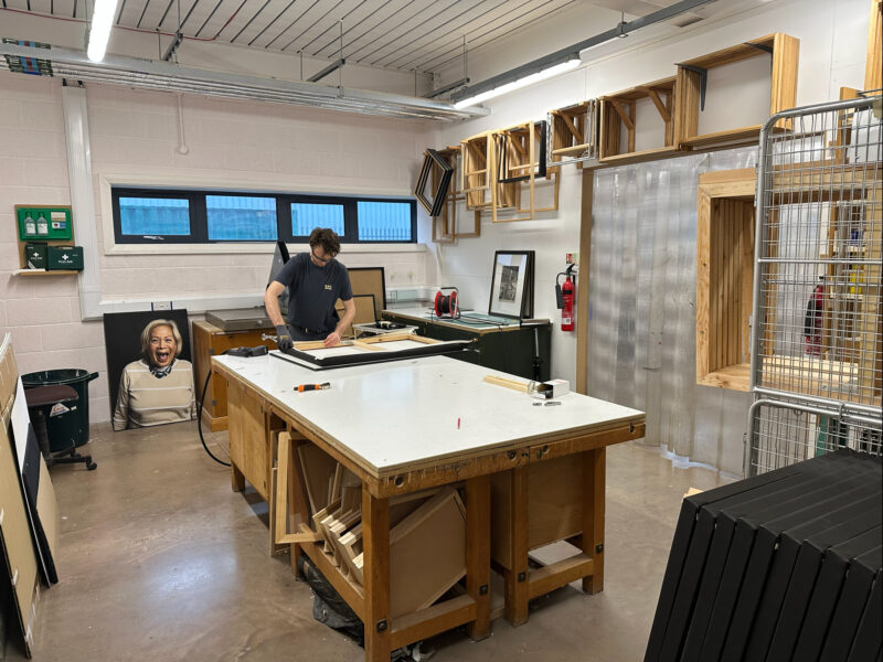 A workshop with wooden frames hanging on the walls and a large centre table. A man leans over the table.