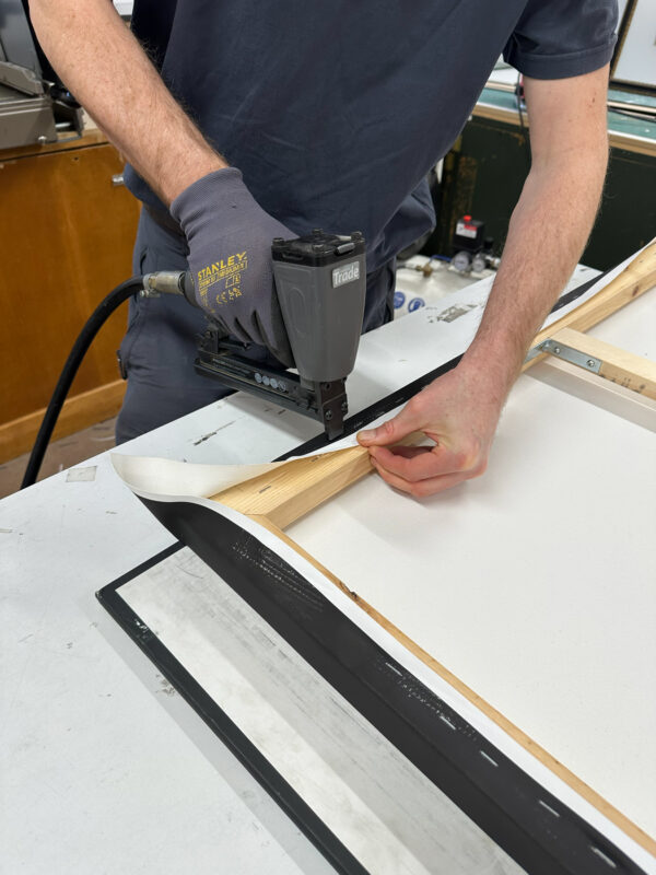 Someone using a nail gun to fix a canvas portrait onto a wooden frame.