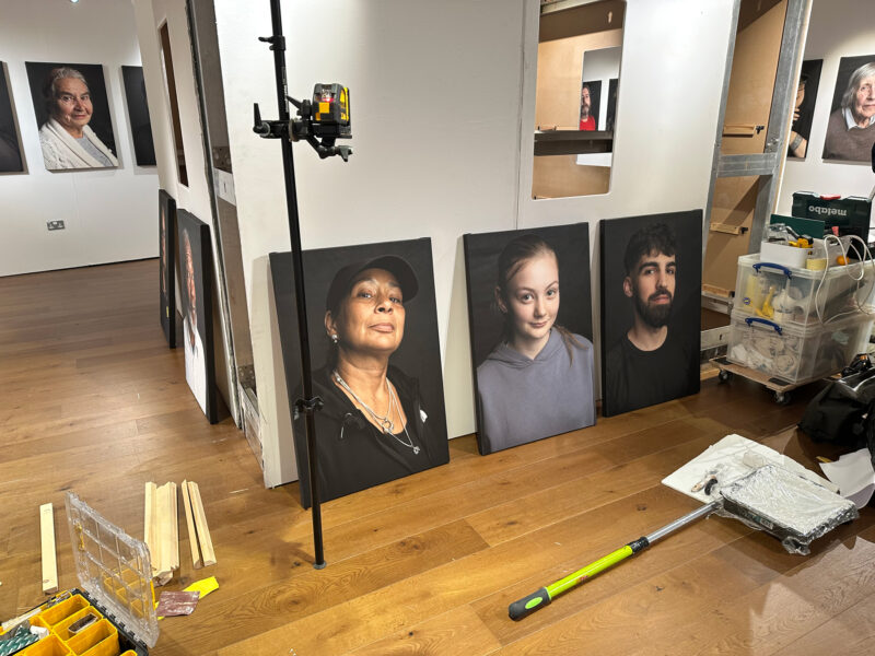 Three canvas portraits resting on the floor, surrounded by decorating materials. More canvases can be seen in the background mounted onto the wall.