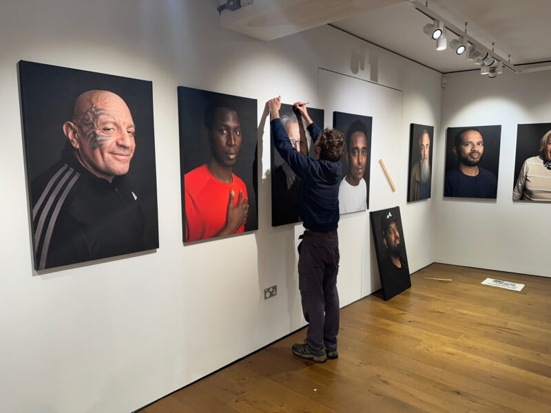 A man hanging large canvas portraits onto a white wall. He is facing away from the camera.