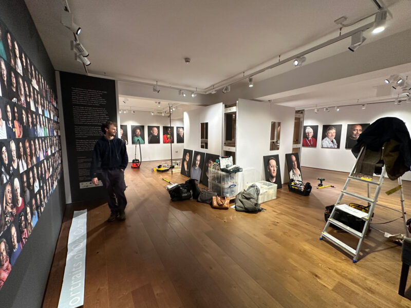 A picture of a gallery in progress. A man stood to the left, surrounded by partially mounted portraits and decorating materials.