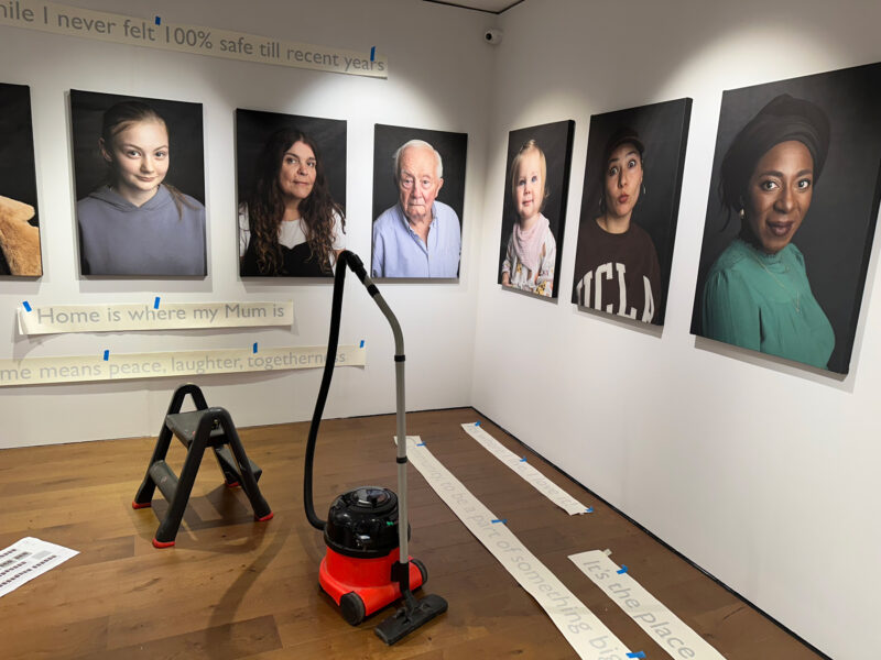 Portraits mounted onto a white wall, with text underneath and a Henry Hoover on the floor.