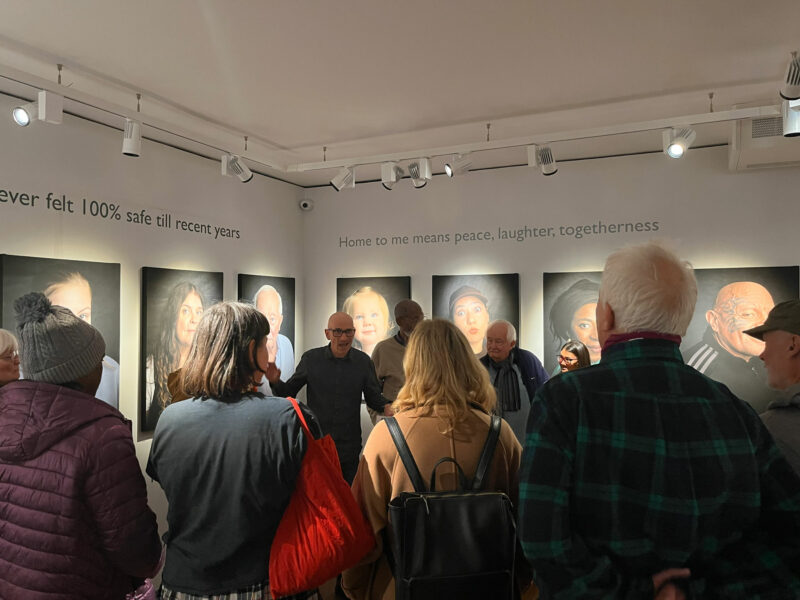A group of people facing away from the camera, listening to a man speak in a gallery of portraits.