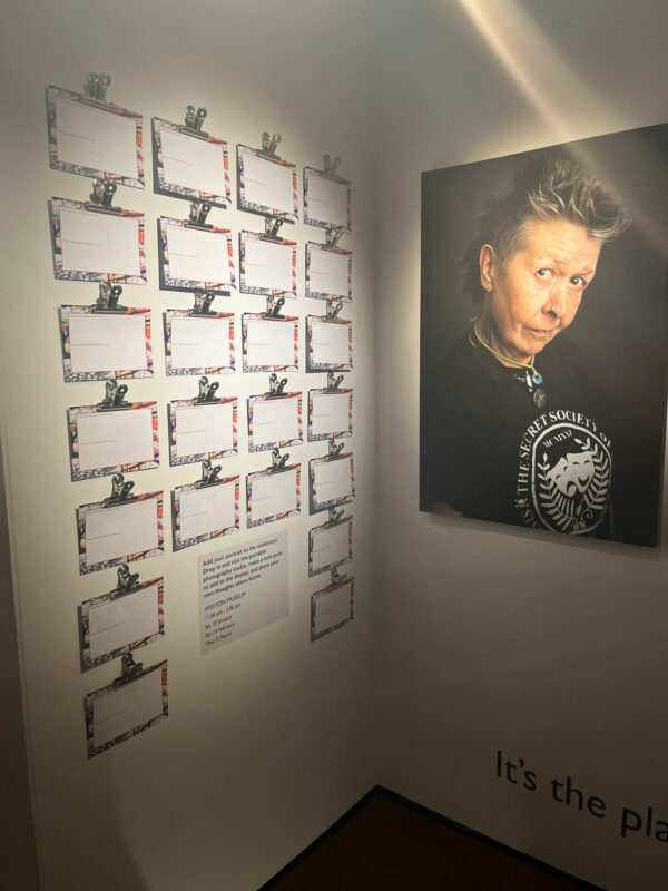 On the right there is a photographic portrait of an older woman in a black t-shirt, on the left there is a wall full of comment cards. These are both mounted in the corner of a gallery.