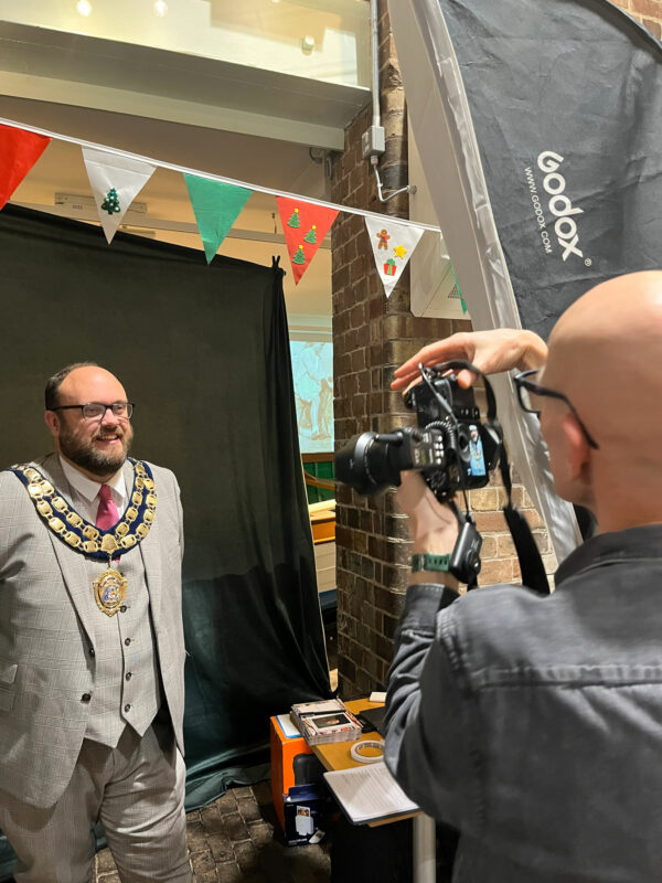 A man taking a portrait photograph of another man, who is wearing a Mayoral outfit. There is some multicoloured bunting over the Mayor's head and he is stood in front of a black background.