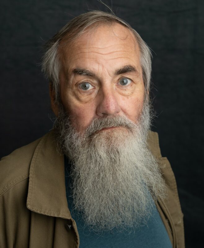 Portrait of an elderly man in a brown coat and blue jumper, with a long grey beard.