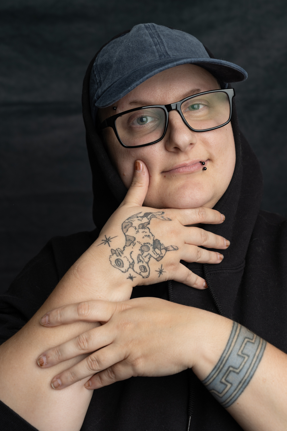 A photographic portrait of a person in a black hoodie and grey baseball cap. They are wearing thick framed black glasses and have a black lip piercing. They have their hands up to their face to show two tattoos.