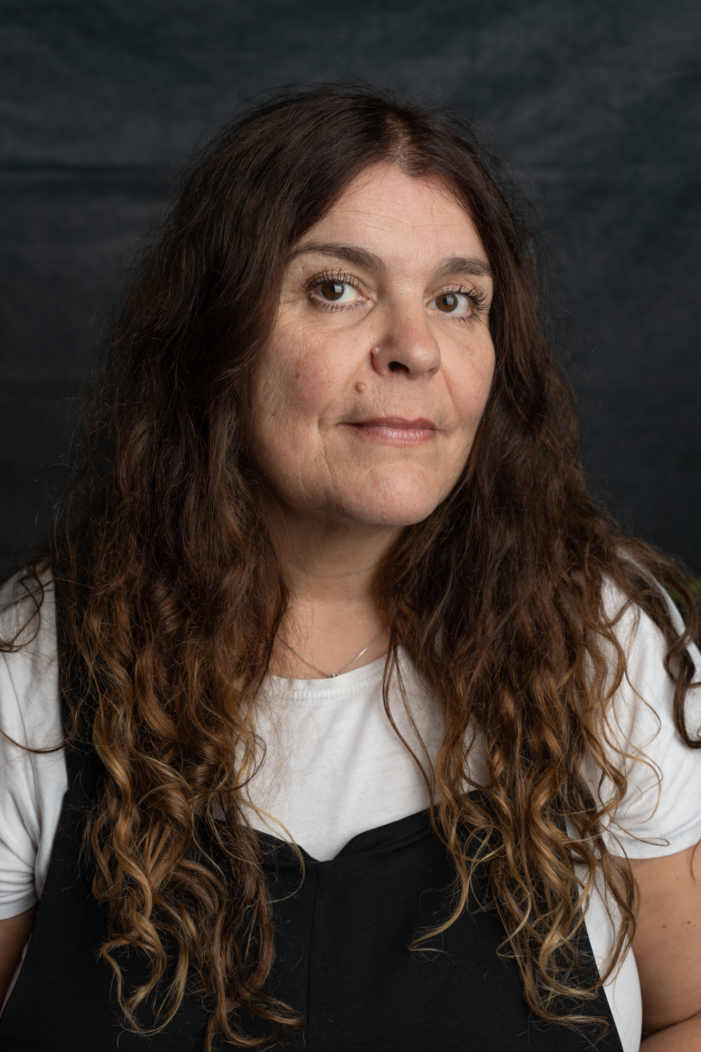 A photographic portrait of a woman with long, curly brown hair. She is not smiling. She is wearing a white t-shirt under black dungarees.