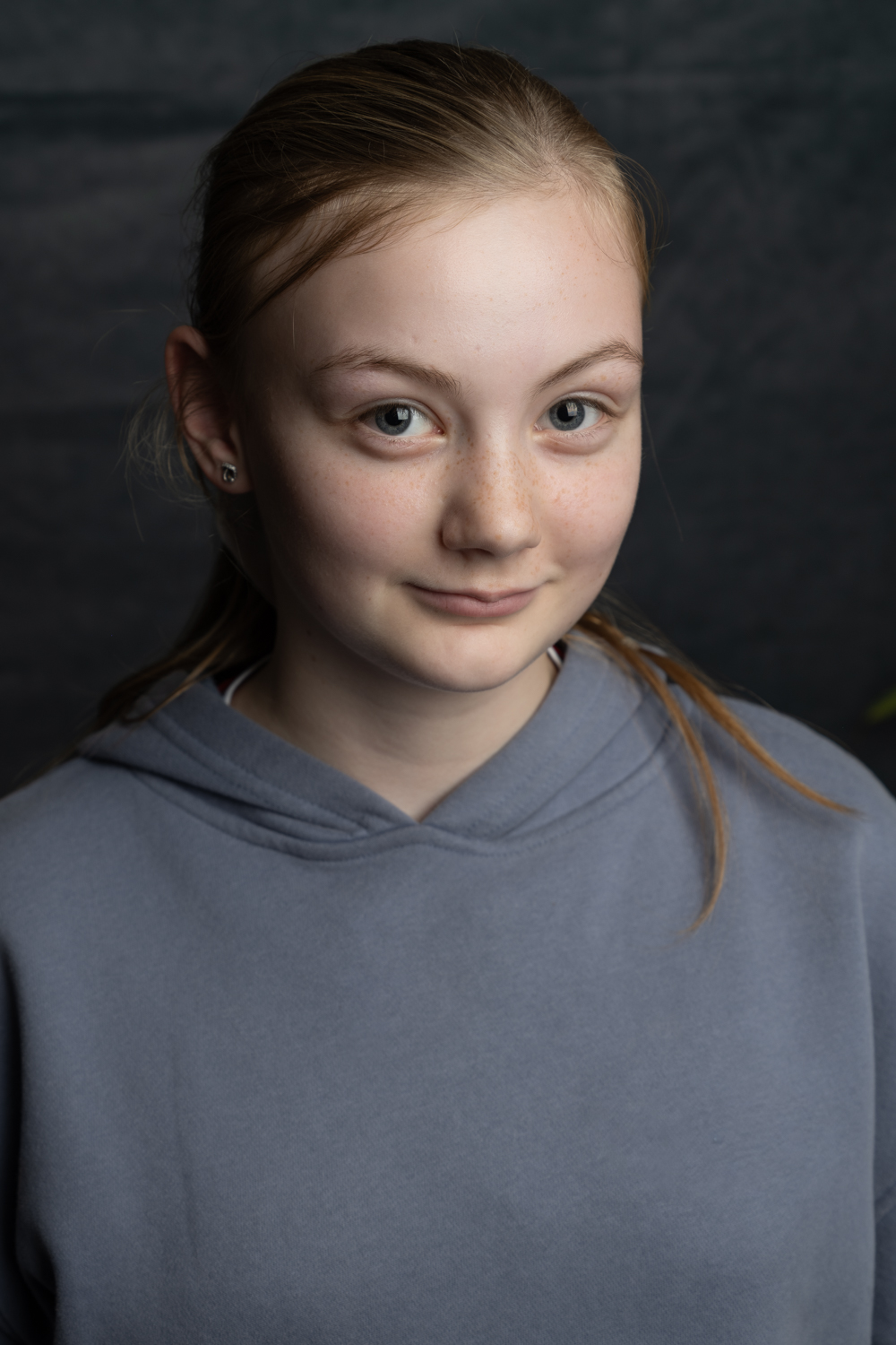 A photographic portrait of a child wearing a grey hoodie. She has long brown hair in a ponytail and is smiling gently.