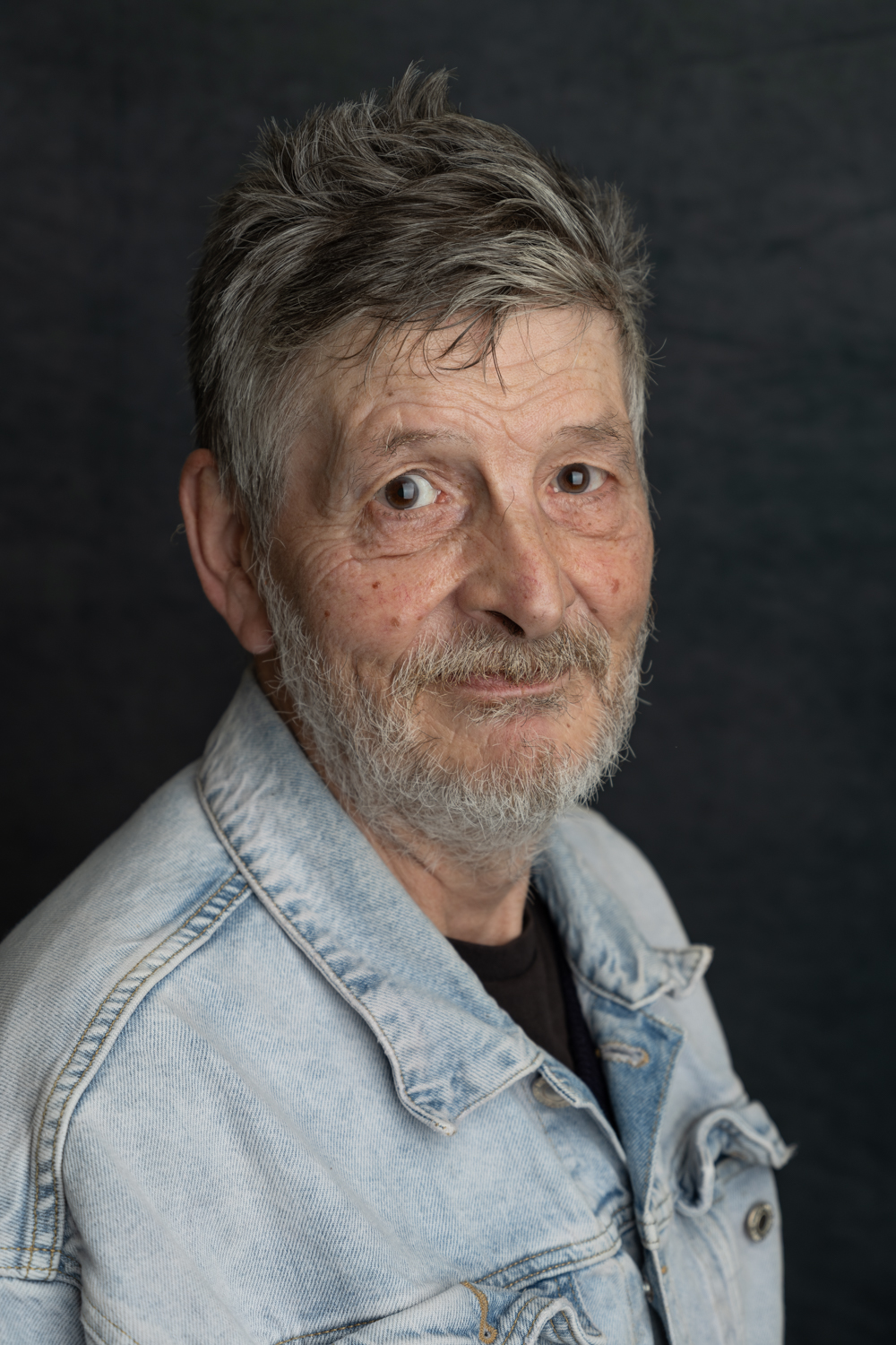 A photographic portrait of a man with grey hair and a grey beard. he is wearing a light blue denim jacket over a black t-shirt.
